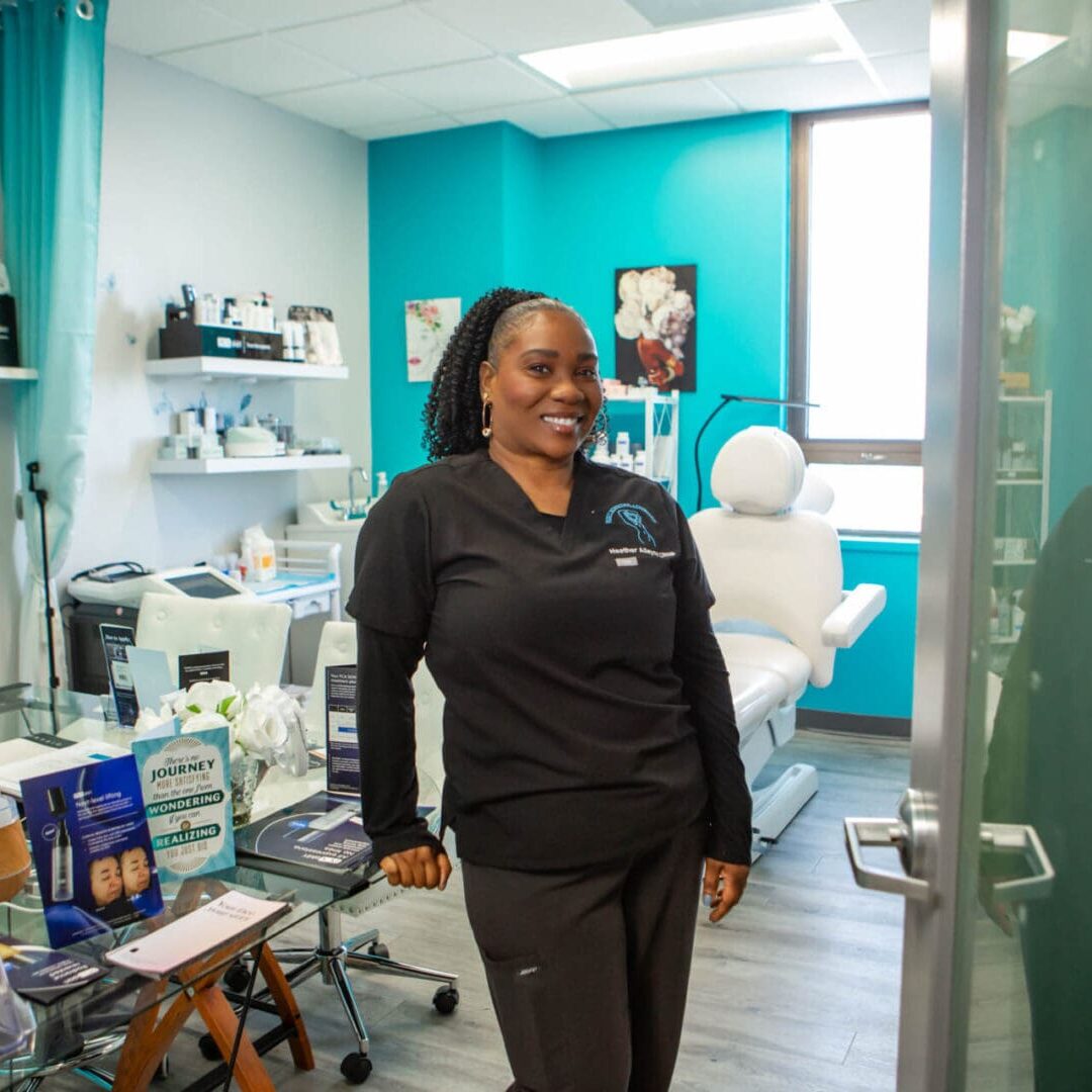 A smiling woman in black scrubs stands in a bright medical room.