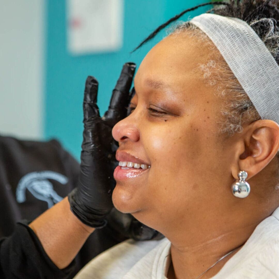 A person wearing black gloves applies makeup to a smiling woman.