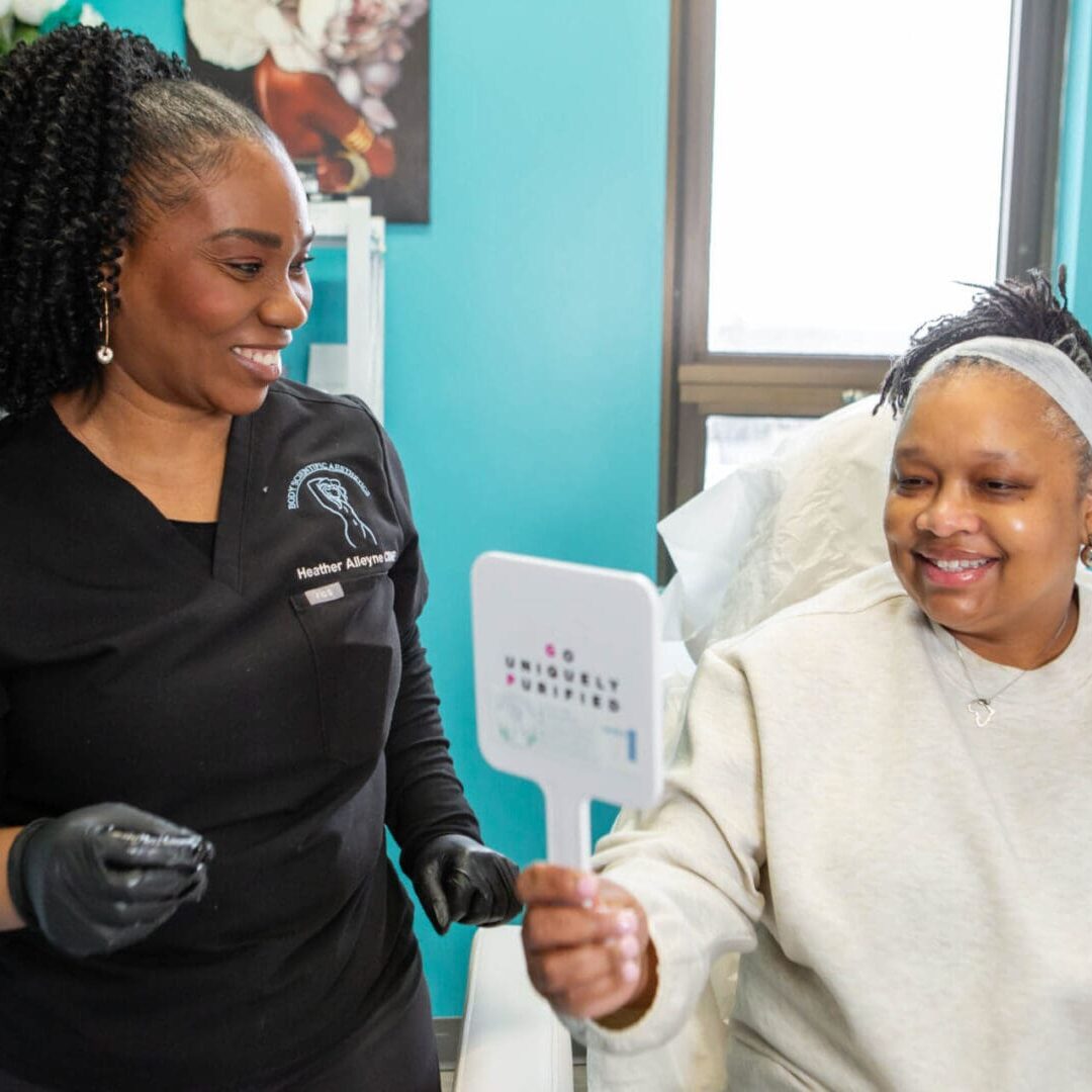 A patient smiles looking at her reflection while a healthcare worker stands nearby.